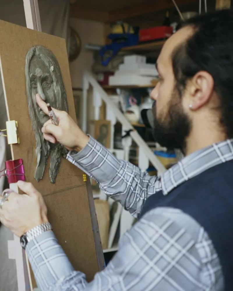 Manos trabajando un rostro modelado en barro sobre un soporte vertical, con una esteca de madera apoyada en la mejilla